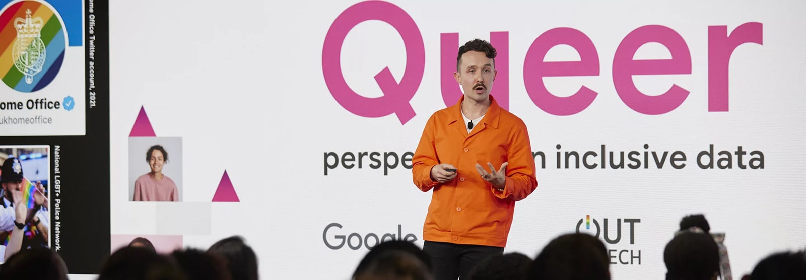 Kevin is standing on stage, he is wearing a bright orange jacket and delivering a talk. In the background is a slide that reads: Queer perspectives on inclusive data.