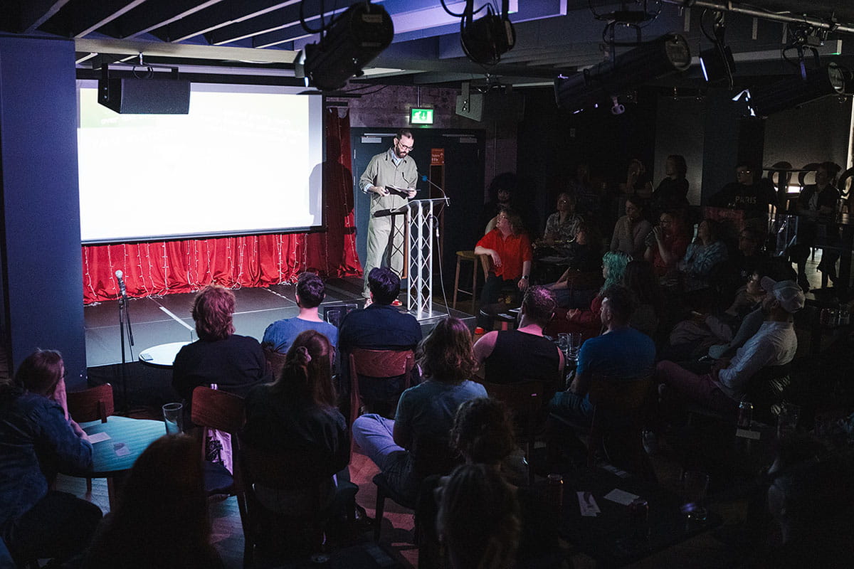 Person presenting on stage in a darkened theatre with the audience watching 